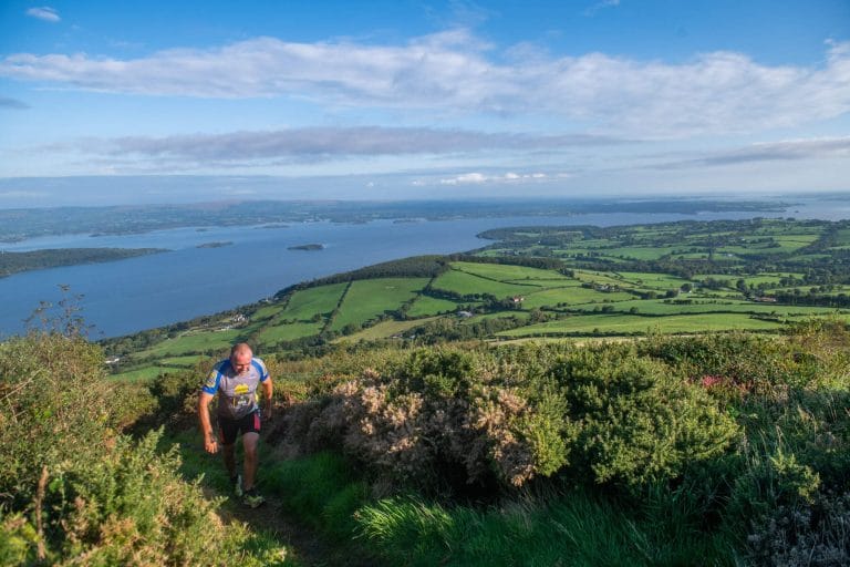 Man hiking uphill on a grassy trail with bushy vegetation, overlooking a scenic landscape of green fields, trees, and a large body of water under a partly cloudy blue sky.