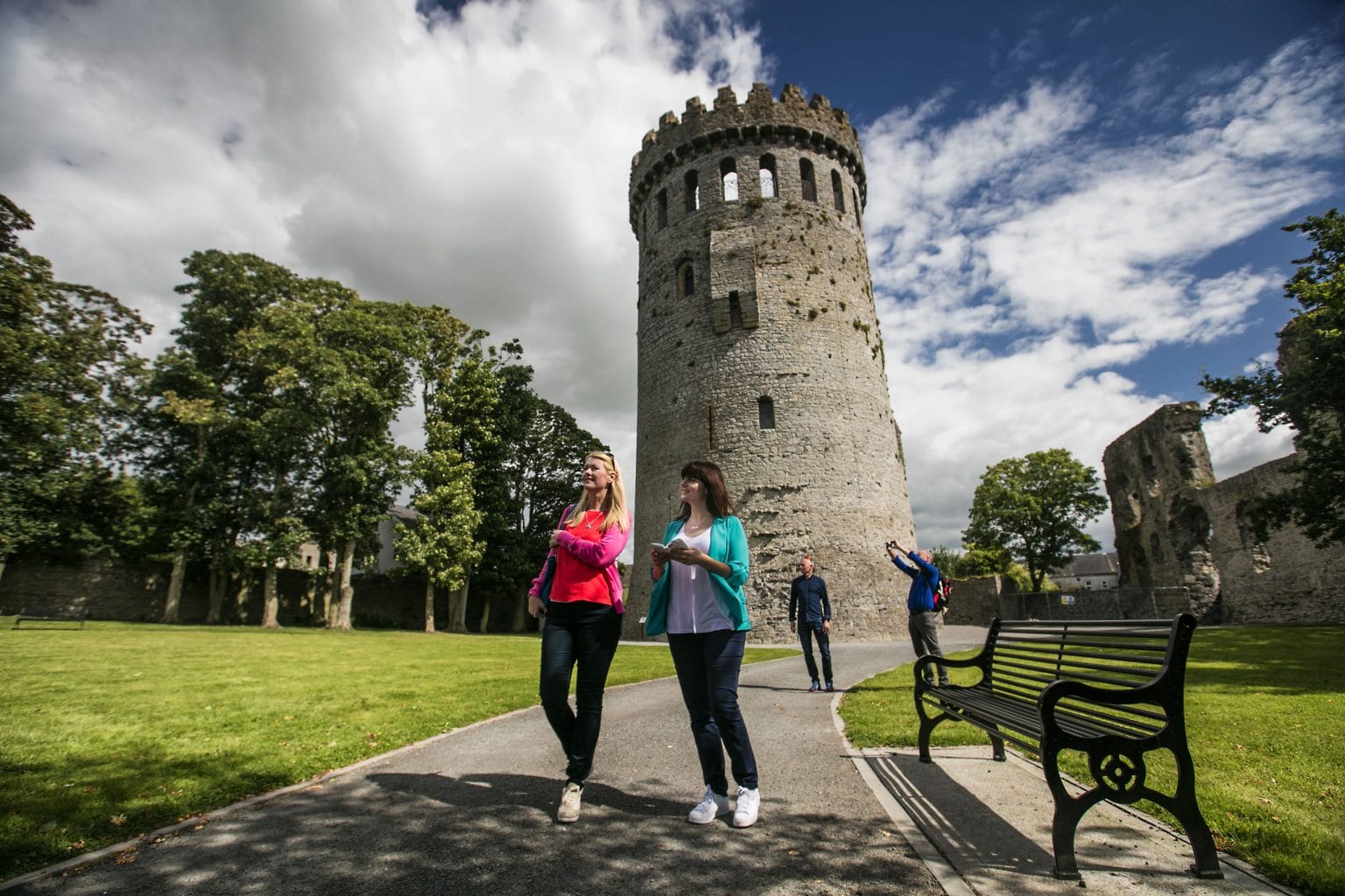 Two women walking and sightseeing near a tall, round medieval stone tower with battlements, surrounded by green grass, trees, and castle ruins; a man in the background is taking a photo, and a black metal bench is on the right side of the paved path.