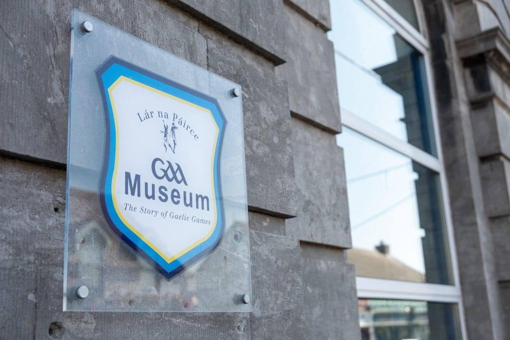 Sign mounted on a stone wall reading "Lár na Páirce GAA Museum The Story of Gaelic Games," with a reflection of a building in the adjacent window.