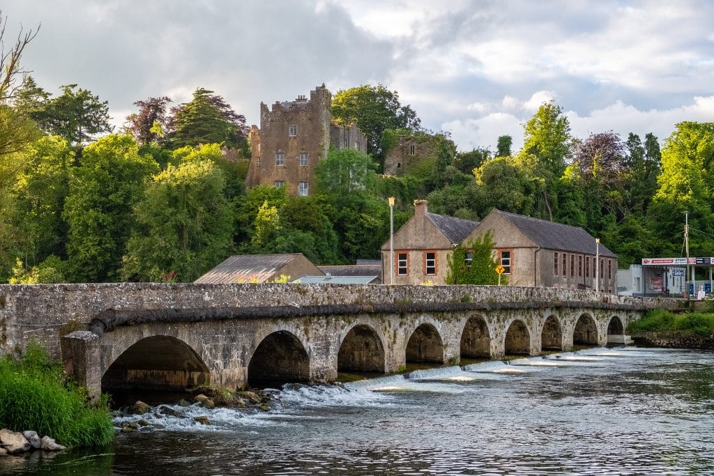Stone arch bridge over a flowing river with a historic castle partially covered by greenery in the background, alongside old stone buildings and lush trees under a cloudy sky.