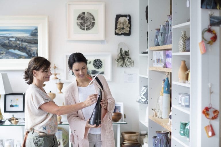 Two women browsing and examining a ceramic decorative item in an art gallery or craft shop, surrounded by various pottery and artwork displayed on shelves and walls.