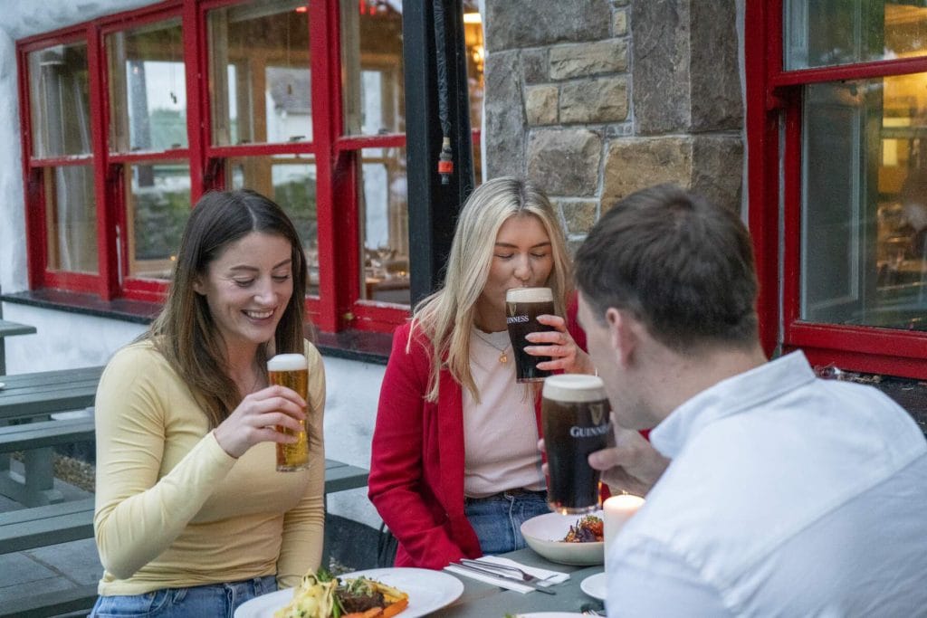 Three people sitting outdoors at a table with food and drinks, two women and one man, enjoying pints of beer; the setting appears to be a pub with red window frames and stone walls.