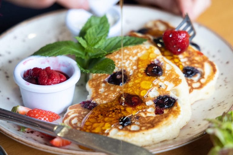 Plate with blueberry pancakes being drizzled with syrup, garnished with fresh mint, a small cup of raspberries, and sliced strawberries, with a hand holding a fork and a berry.