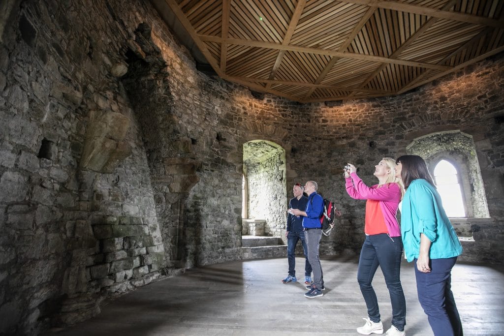 Four adults touring the interior of an ancient stone building with arched windows, admiring the structure and taking photographs under a wooden coffered ceiling.