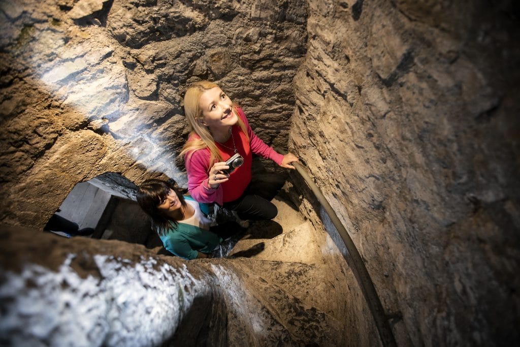 Two women ascending a narrow stone spiral staircase inside a historic or ancient structure, one holding a camera and looking upwards.